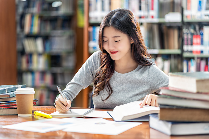 Asian young Student in casual suit reading and doing homework in library of university or colleage with various book and stationary on the wooden table over the book shelf background, Back to school